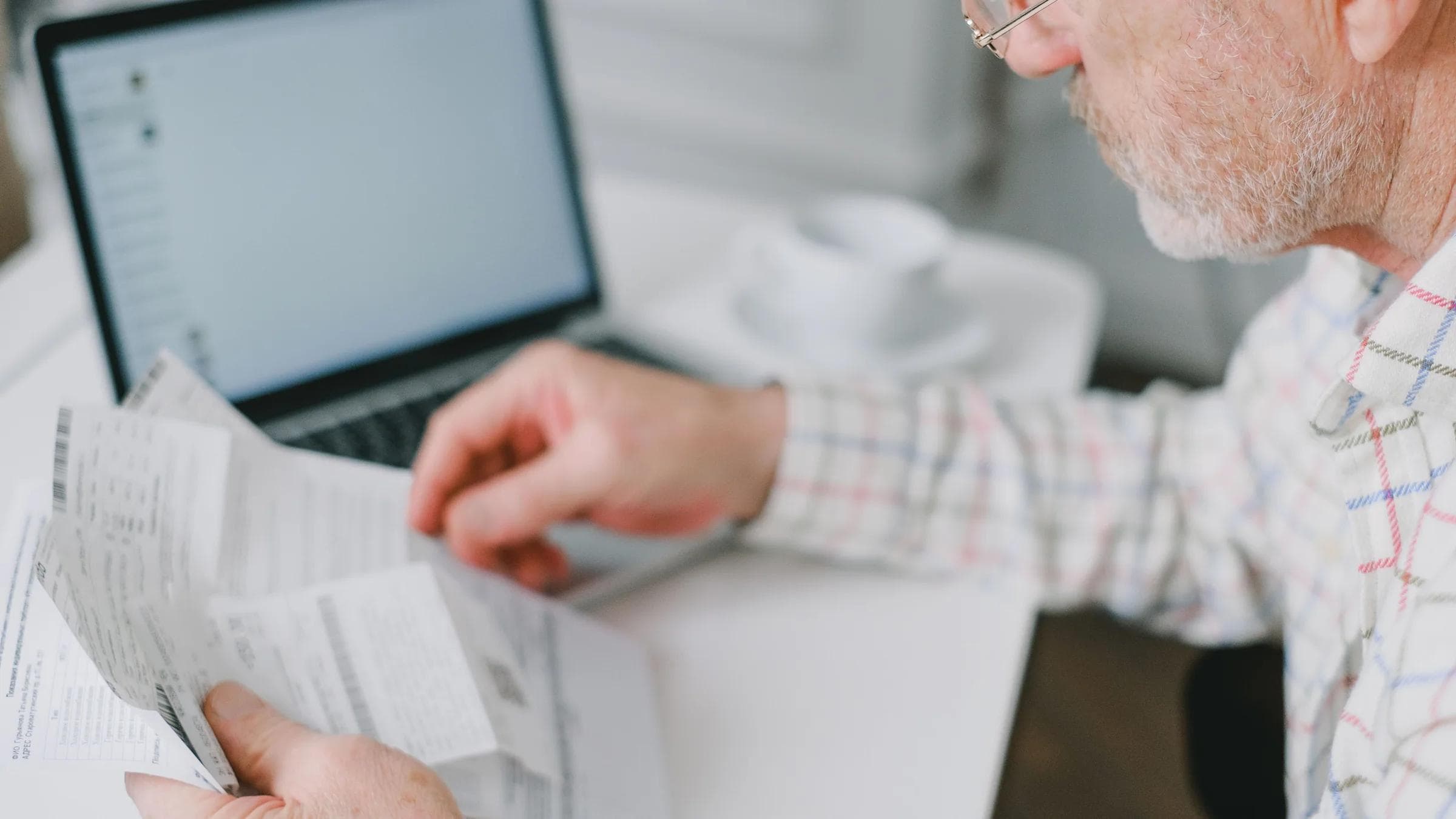 Elderly man wearing eyeglasses reading documents next to a laptop at home.