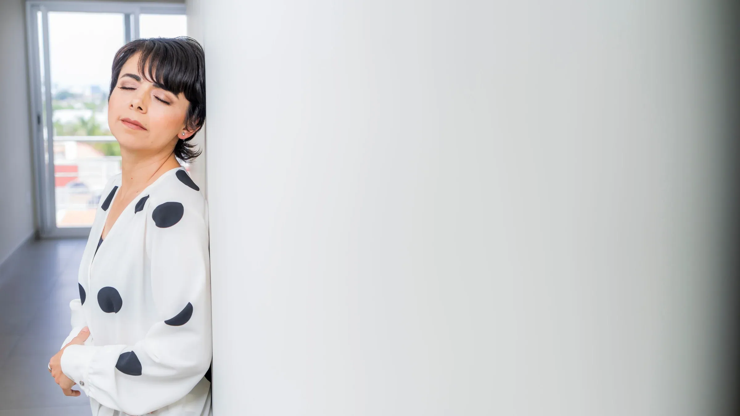 Adult woman in a stylish polka dot blouse with eyes closed, leaning against a white wall indoors.