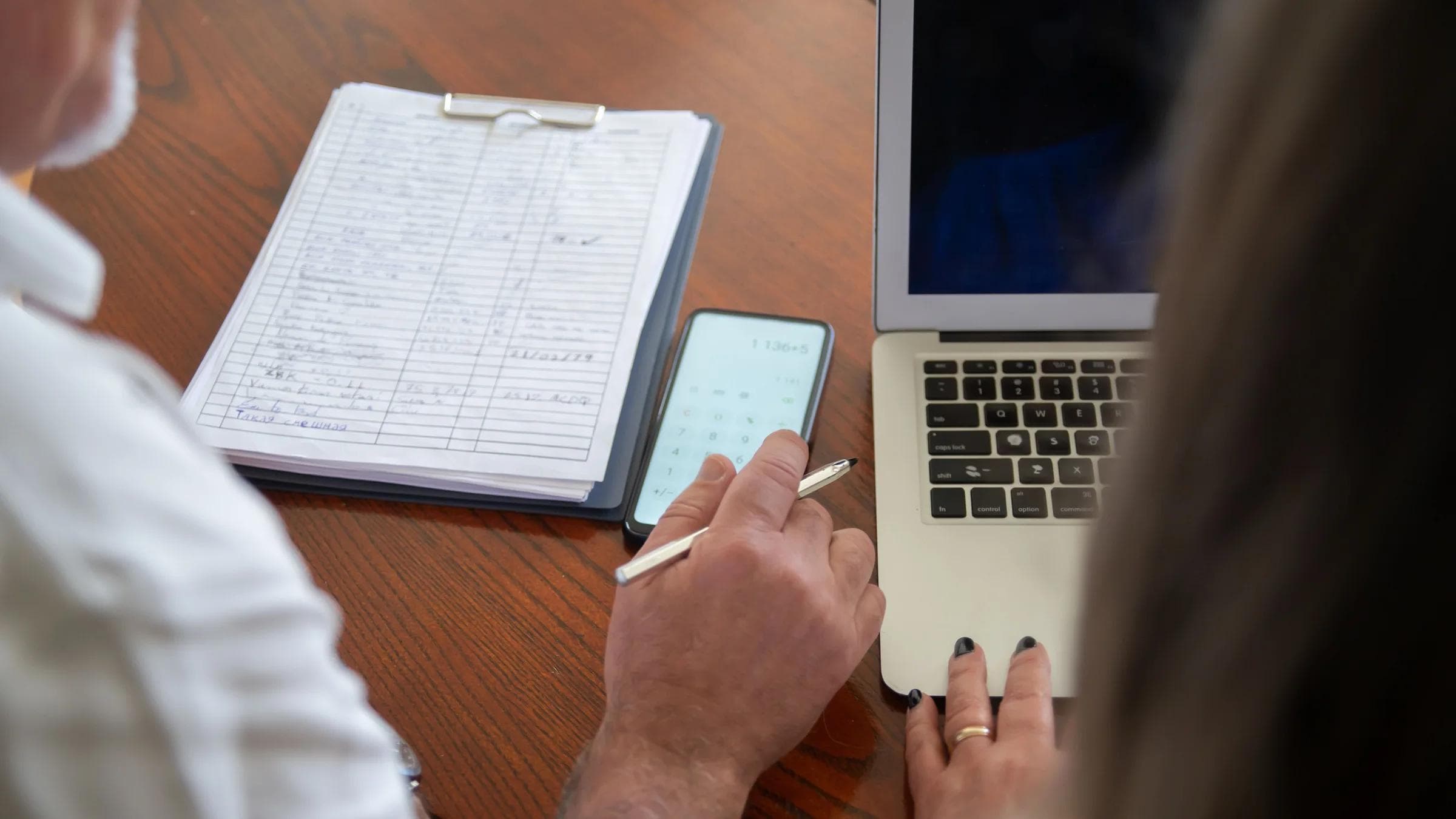 Two adults working at a desk with a laptop, smartphone, and documents. Collaborative workspace scene.