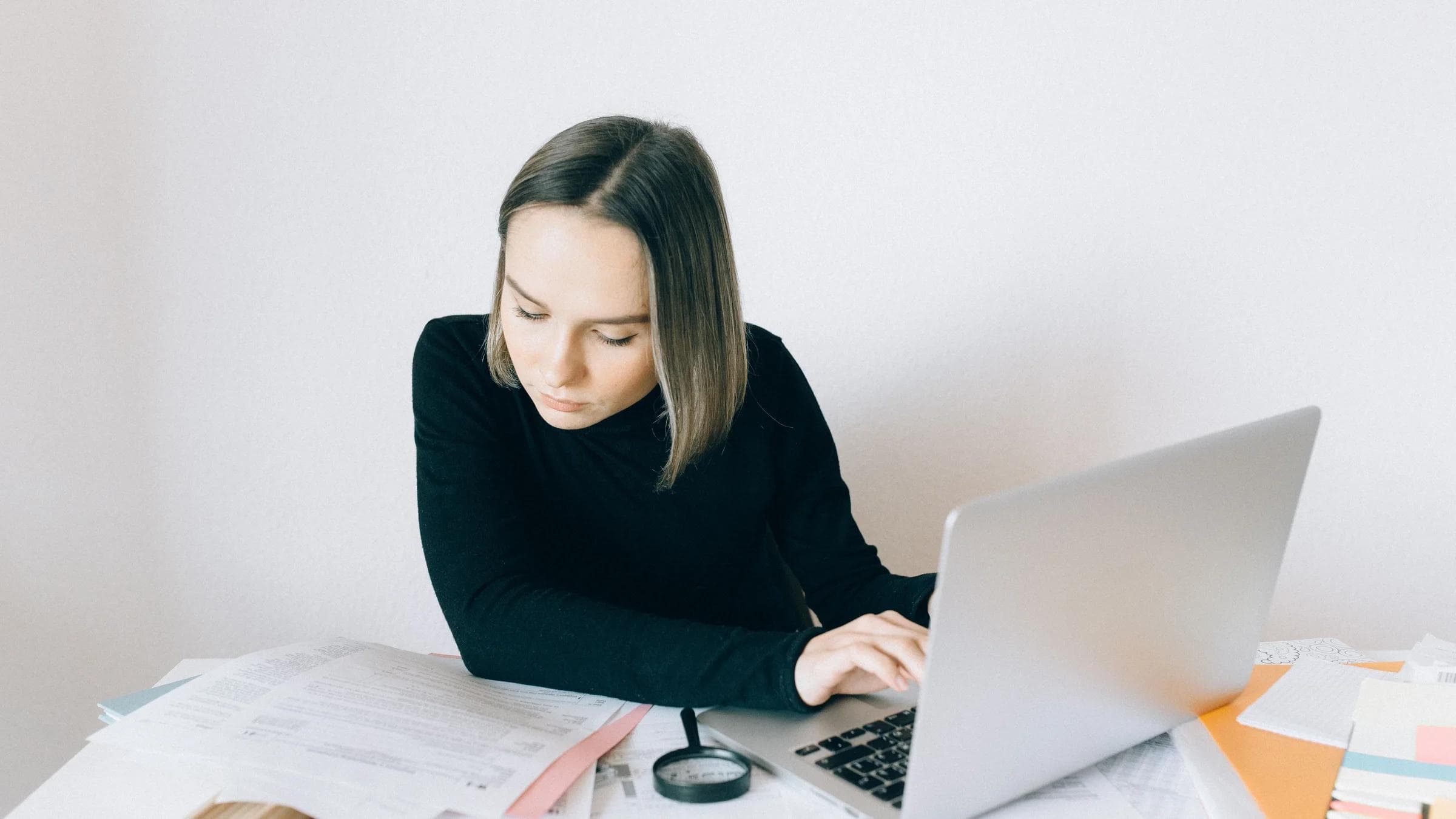 Focused woman researching on a laptop, surrounded by paperwork in a modern office.