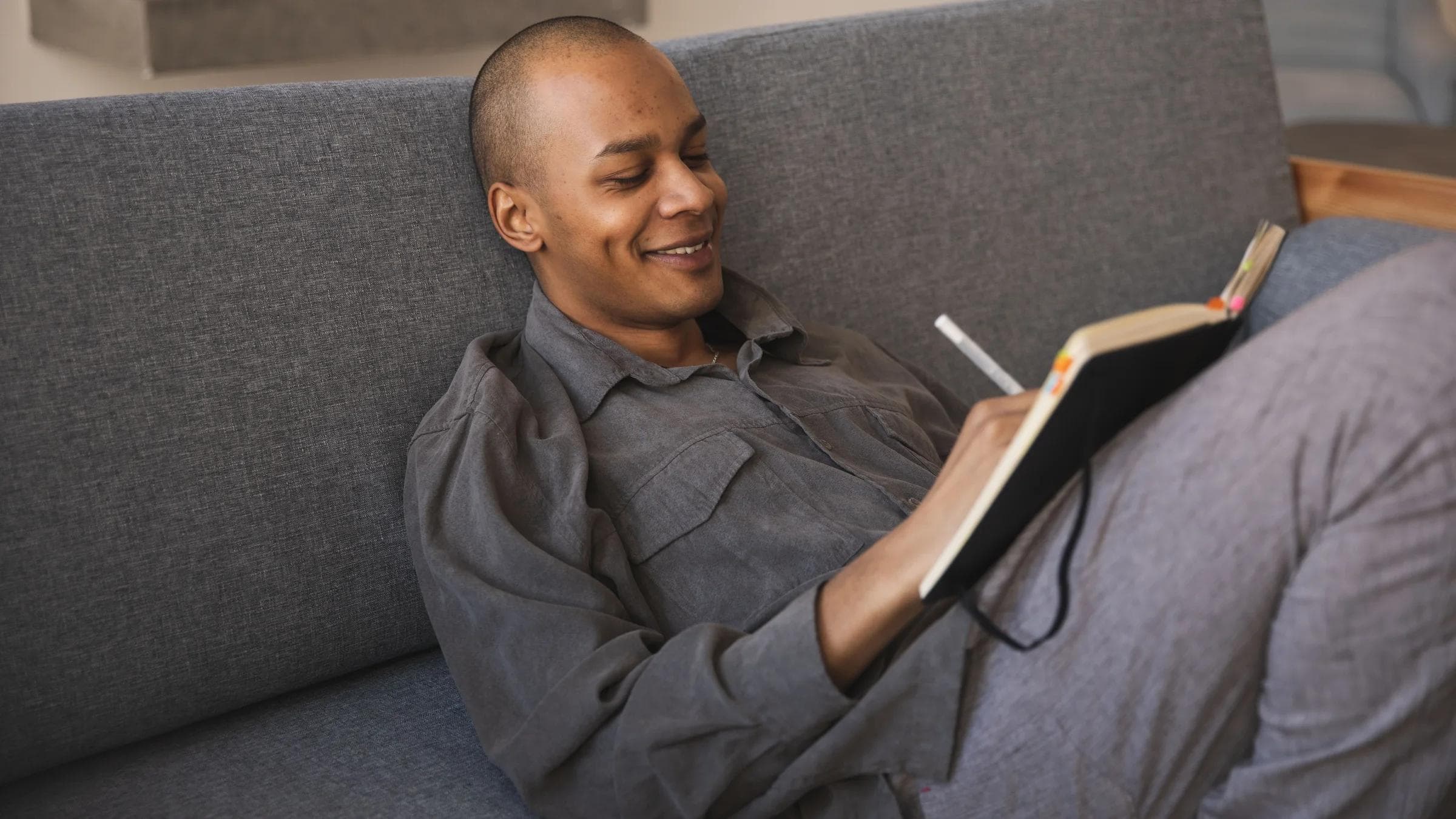 A man smiles while writing in a journal on a comfortable sofa, enjoying a moment of relaxation.