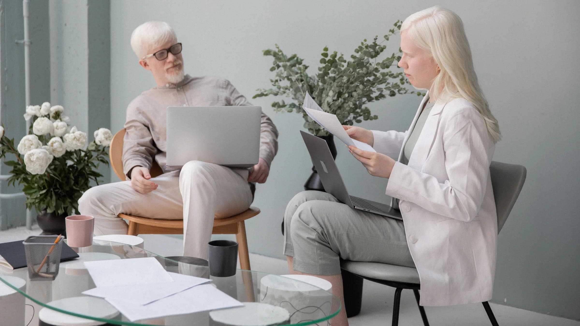 Focused woman with documents sitting against wall in office with table and flowers near man with white beard and laptop while working.